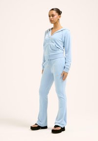 Šviesiai mėlynas zip-up džemperis su atitinkančiomis kelnėmis, pagamintas iš minkštos medžiagos. Yra priekinės kišenės, gobtuvas ir subtilūs auksiniai akcentai. Platėjantys kelnių šonai.