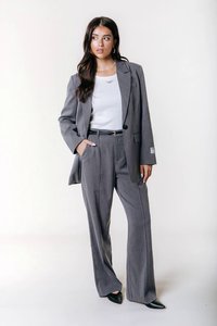 Γυναίκα που φοράει γκρι ρίγες κοστούμι με παντελόνι φαρδύ, άσπρο τοπ, μαύρη ζώνη και μαύρες μυτερές γόβες, στέκεται μπροστά σε άσπρο φόντο.