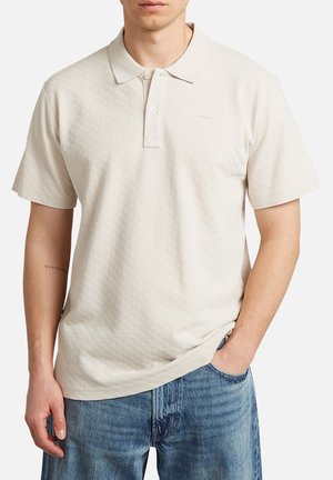 Mann trägt ein hellbeiges kurzärmeliges Poloshirt und blaue Jeans, mit einer Hand in der Tasche und sichtbarem Unterarmtattoo.