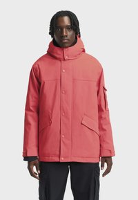 Vyras, vilkintis koralų raudoną žieminę striukę su užsegamais kišenėmis ir juodomis cargo kelnėmis, stovi priešais baltą fono.