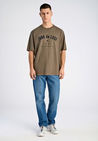 Camiseta verde oliva con "JUNK de LUXE" impreso en azul marino, combinada con jeans de denim azul y zapatillas blancas. Diseño sencillo y de corte relajado.