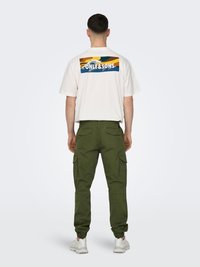 Camiseta blanca con diseño gráfico colorido en la espalda, combinada con pantalones cargo verdes con bolsillos laterales y puños elásticos. Se ven zapatillas blancas.