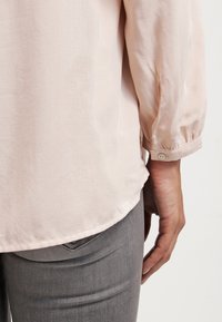 Blouse rose clair à manches longues légèrement bouffantes, texture lisse, avec détail de bouton aux poignets, associée à un jean gris.