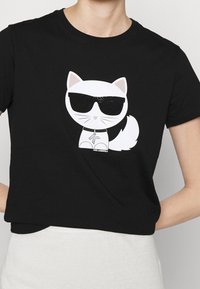 Černé bavlněné tričko s grafickým motivem bílé kočky se slunečními brýlemi, hravý design a malý textový detail "KARL LAGERFELD" ve spodní části.