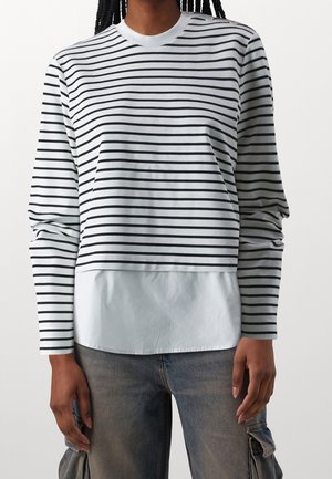 Persona que lleva una camiseta de manga larga con rayas horizontales negras y blancas sobre una camiseta blanca, combinada con jeans cargo azul deslavado.