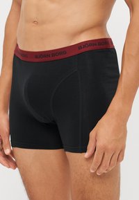 Schwarze Boxer-Brief mit einem burgunderroten elastischen Bund, auf dem "BJÖRN BORG" in schwarzer Schrift steht. Glatter Stoff mit enganliegendem Design und Nähdetails.