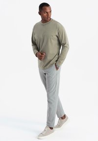 Mann in olivgrünem Langarmshirt, hellgrauen Hosen und beigen Sneakern, der zur Seite schaut, mit einer Hand in der Tasche vor weißem Hintergrund.