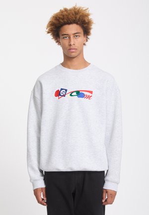 Jeune homme aux cheveux bouclés portant un sweat-shirt gris clair avec un graphisme abstrait coloré et un pantalon noir sur fond blanc.