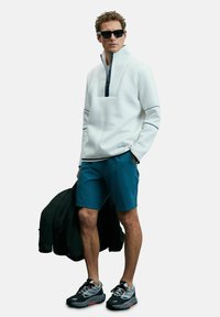 Homme portant un pull blanc, un short bleu, des lunettes de soleil noires et des baskets, tenant une veste noire dans une main.
