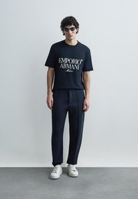 T-shirt de manga curta azul-marinho com logótipo branco "Emporio Armani", combinado com calças escuras e ténis brancos, vestido por um modelo masculino.