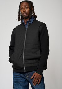 Veste noire zippée avec un panneau avant matelassé, des poignets côtelés et une texture lisse. Associée à une chemise en denim bleu en dessous.