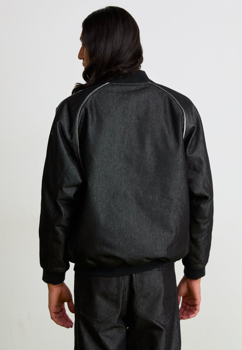 Schwarze Bomberjacke mit strukturierter Oberfläche, elastischen Bündchen und Saum sowie dezenten grauen Nähdetails. Kombiniert mit passenden Hosen.