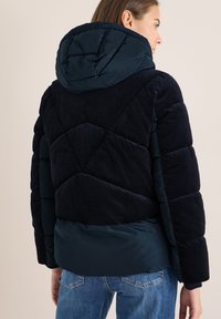 Veste matelassée bleu marine avec un dessus en velours côtelé, design matelassé et capuche. Dotée d'une partie inférieure lisse et de poignets élastiques.