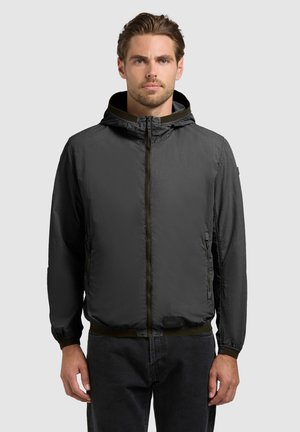 Egy férfi sötétszürke, kapucnis cipzáras dzsekit visel, és egy egyszerű, világosszürke háttér előtt áll, elölről nézve.