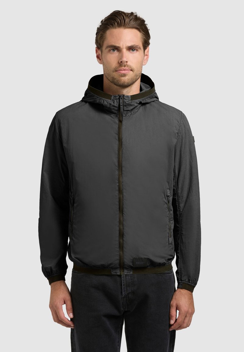 Muž oblečený v tmavosivej vetrovke so zipsom a kapucňou, stojaci oproti jednoliatemu svetlosivému pozadiu, tvárou vpred.