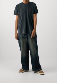 Černé tričko s krátkým rukávem a kulatým výstřihem, se červeným logem na levé části hrudi, spárované s volnými světlými modrými džíny a hnědými teniskami.