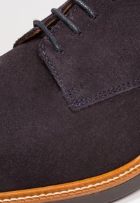 Chaussure en daim bleu marine foncé avec une semelle en cuir beige, dotée de lacets foncés et de surpiqûres contrastantes, mettant en valeur une texture lisse et un design poli.