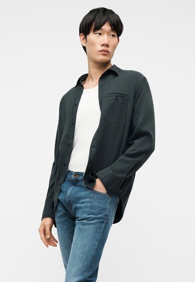 Νεαρός άνδρας που φορά σκούρο πράσινο πουκάμισο με κουμπιά πάνω από λευκό μπλουζάκι και μπλε τζιν, στέκεται με το ένα του χέρι στην τσέπη, κοιτάζοντας προς το πλάι.