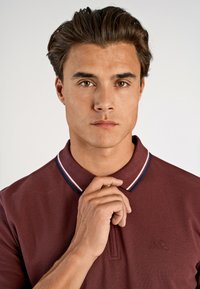 Burgundowa koszulka polo z teksturowanej tkaniny, z trzema guzikami i kontrastowym kołnierzykiem w paski w kolorze granatowym i białym.