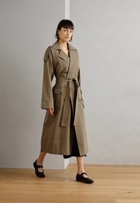 Cappotto trench khaki con vita cinturata, due grandi tasche frontali e dettagli con bottoni. Indossato sopra un vestito nero, abbinato a scarpe nere.