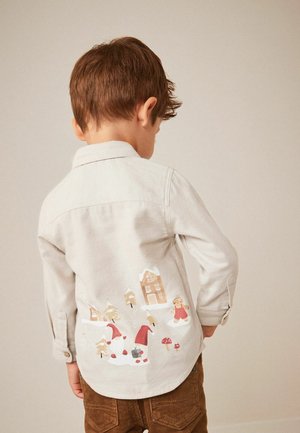 Enfant vu de dos portant une chemise beige avec un motif de village hivernal et de gnome, associée à un pantalon en velours côtelé marron, sur fond uni.