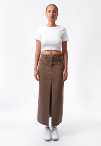 Falda maxi de mezclilla marrón con una abertura frontal, combinada con una camiseta ajustada de manga corta blanca. La modelo lleva zapatillas blancas y está de pie frente a un fondo plano.
