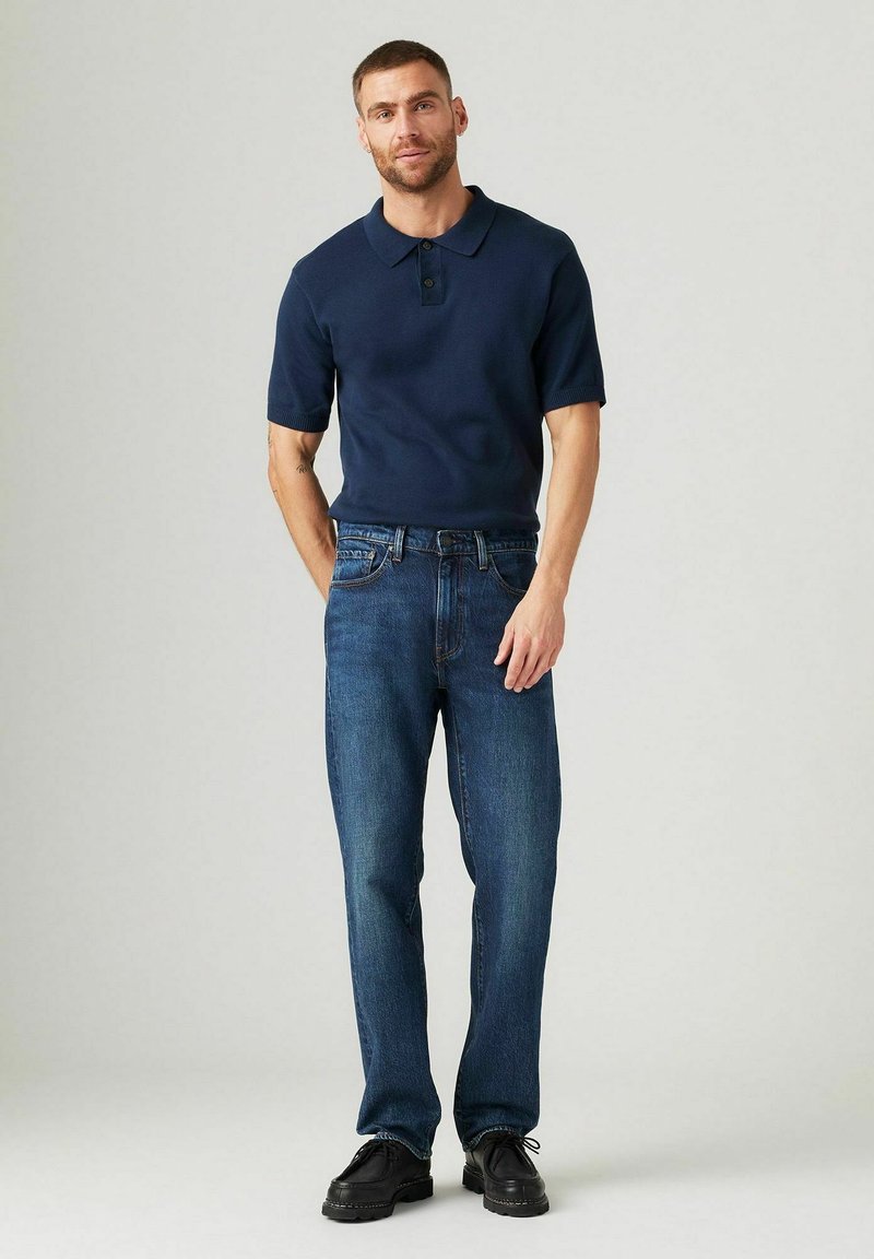 Marineblaues Polohemd mit kurzen Ärmeln und Kragen, kombiniert mit dunkelblauen geraden Jeans und schwarzen Schnürschuhen. Minimale Details.