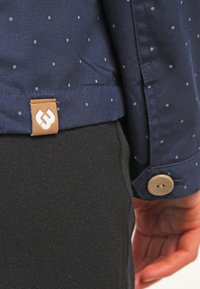 Marinblus med små vita prickar, prydd med en läderetikett och en rund metallknapp. Blusen är kombinerad med åtsittande svarta byxor.