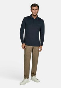 Navy longsleeve poloshirt met een borstzak, gecombineerd met tan tapered broeken en witte sneakers op een effen achtergrond.