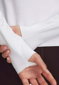 Fehér, hosszúujjú ing, testhez simuló dizájnnal, hüvelykujjlyukakkal a mandzsettákon, sima szövettextúrával és minimális varrási részletekkel.