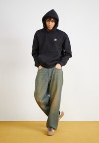 Μαύρη κουκούλα με τσέπη καγκουρό, χαλαρά ξεθωριασμένα μπλε τζιν και ανοιχτό καφέ αθλητικά παπούτσια. Η κουκούλα έχει λευκό λογότυπο στο στήθος.