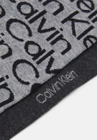 Sivý textil s čiernym písmom, s názvom značky "Calvin Klein" v rôznych veľkostiach. Hladká textúra s kontrastnými vzormi.