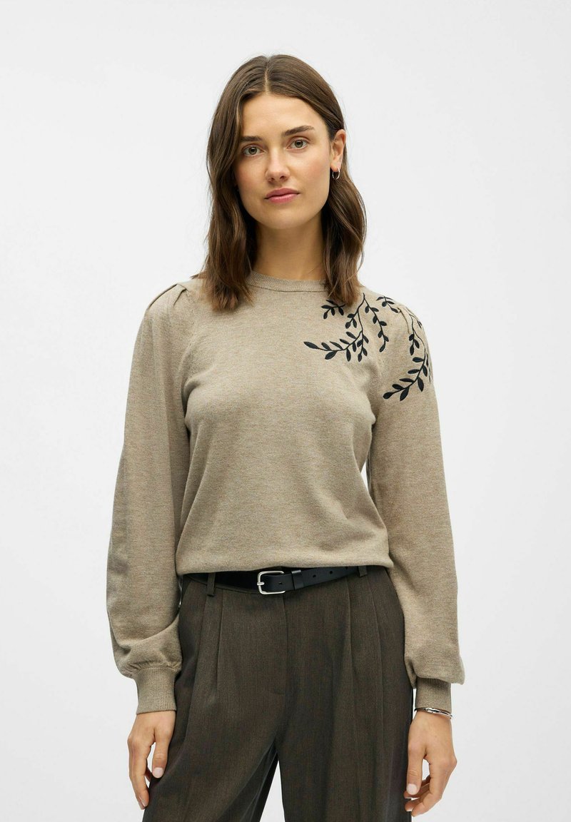 Beige Pullover mit langen Ärmeln, verziert mit schwarzer Blattstickerei an einer Schulter und einem gerippten Rundhalsausschnitt. Kombiniert mit hochtaillierten braunen Hosen.