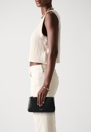 Black leather clutch with gold hardware, featuring a zip closure. Model wears a cream knitted crop top and light-colored jeans.