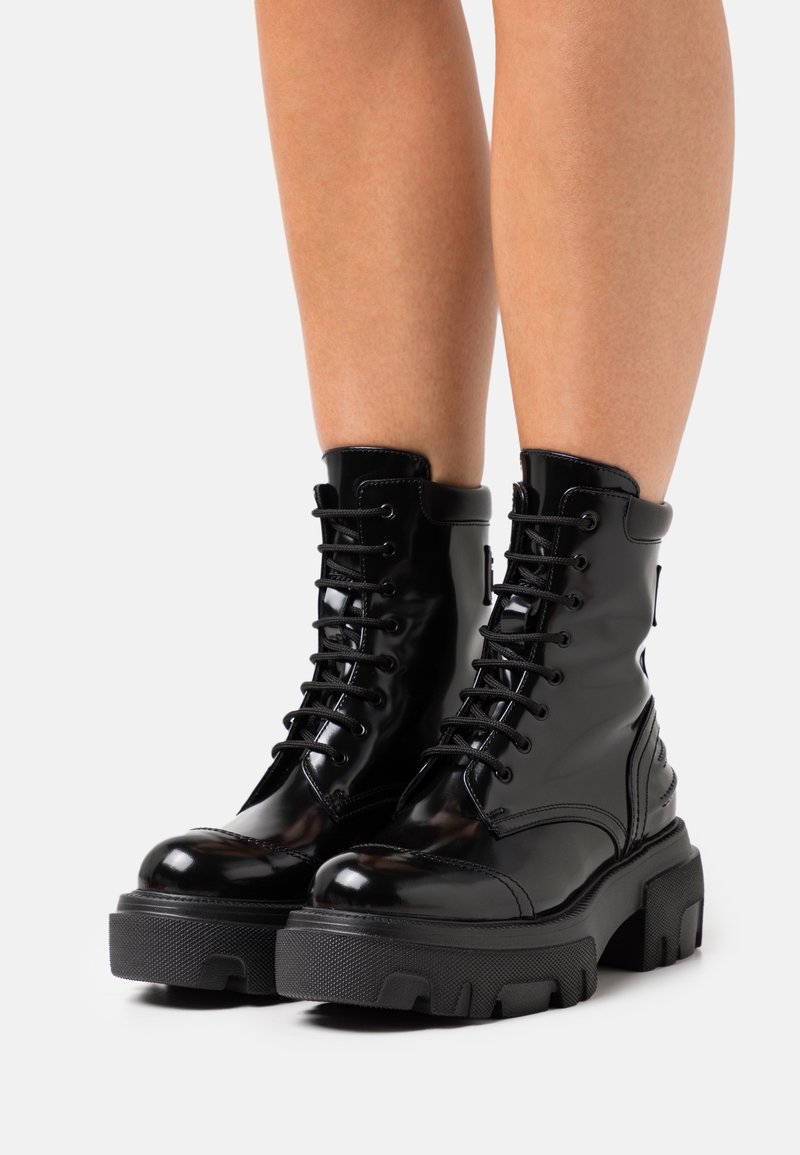 Bottes de combat noires brillantes à lacets avec semelles épaisses et robustes portées sur des jambes nues sur un fond clair uni.