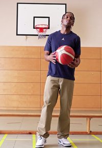 Basketballspiller i en blå skjorte med et logo holder en rød og hvid basketball. Han har beige bukser og hvide sneakers på. Gymnasiebaggrund.