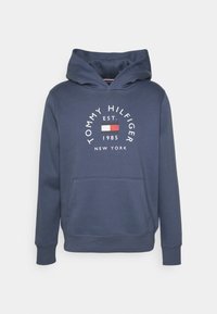 Felpa blu navy con tasca frontale, caratterizzata da un logo circolare "Tommy Hilfiger" e stampa dell'anno di fondazione in bianco. Tessuto morbido con polsini a coste.