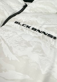 Witte pufferjack met een subtiel textuurpatroon, voorzien van een zwarte rits en het "BLCK BNNS" logo in vetgedrukte zwarte letters.