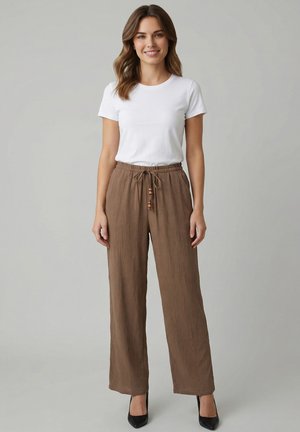 Donna sorridente con capelli alla lunghezza delle spalle, indossa una camicia bianca a maniche corte, pantaloni marroni con coulisse e tacchi neri, in piedi di fronte a uno sfondo grigio.