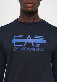 Jūras spēku puloveris ar lielu zilu Emporio Armani logo priekšpusē. Apaļa kakla izgriezuma un teksturēta auduma ar gludu apdari.