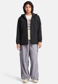 Czarna kurtka z kapturem, z elastycznymi mankietami i zamkiem z przodu, noszona na białym T-shircie z grafiką oraz luźnych, jasnoszarych spodniach typu cargo.