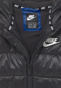 Czarna kamizelka puchowa z pikowanym wzorem, nylonowy zewnętrzny materiał, polarowe podszewki, zamek błyskawiczny z przodu oraz wyraźne białe logo Nike na klatce piersiowej i taśmie na szyi.