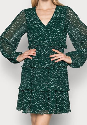 Zielona, warstwowa sukienka z dekoltem w kształcie litery V i długimi, przezroczystymi rękawami. Wzór w groszki oraz elastyczny, falbaniasty pas w talii.