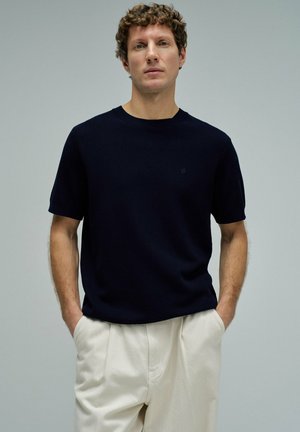 Hombre con cabello rizado que lleva una camisa azul marino de manga corta y pantalones plisados crema, de pie con las manos en los bolsillos contra un fondo liso.