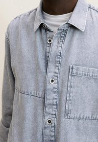 Camicia in denim di colore azzurro chiaro con colletto abbottonato, tasca sul petto e bottoni in metallo. Presenta una vestibilità comoda e una leggera texture.
