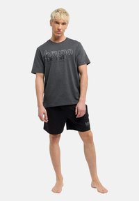 Donkergrijze katoenen t-shirt met vetgedrukte witte tekst en zwarte shorts met logodetail. Model staat blootsvoets, met ontspannen armen langs de zij.