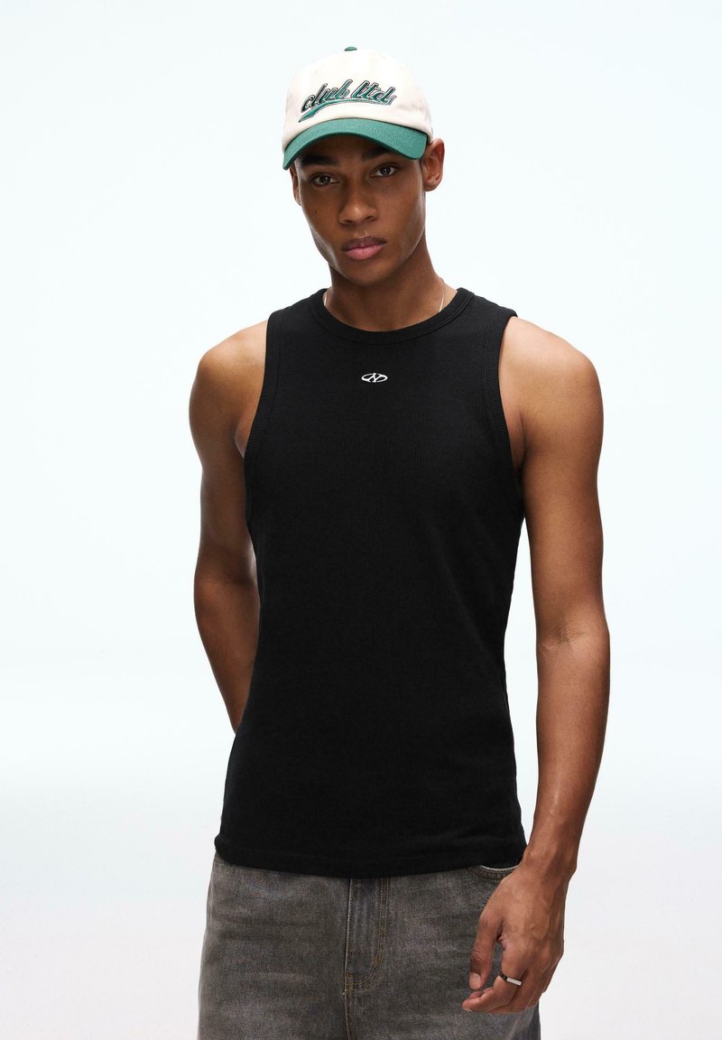 Mladić u crnoj majici bez rukava, sivim trapericama i bež-zelenoj kačket kapi stoji ispred jednostavne bijele pozadine.