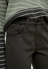 Jean noir en denim avec une texture lisse, doté d'un bouton de fermeture, de poches avant et de détails de couture subtils. Porté sous un pull rayé.