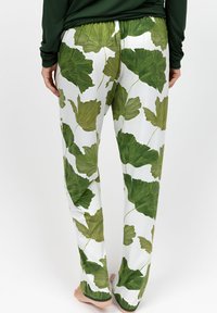 Persoon in witte broek met grote groene bloemenprint, staande op blote voeten tegen een effen achtergrond, van achteraf bekeken.