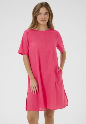 Kobieta ubrana w jasnoróżową, krótkorękawową sukienkę do kolan z bocznymi rozcięciami, stojąca z jedną ręką w kieszeni na jednolitym tle.